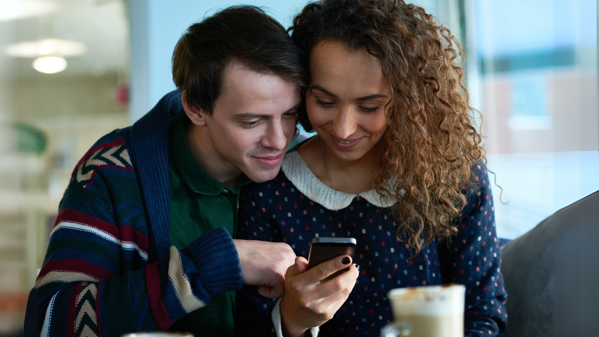 A couple sitting together in a cafe, smiling while looking at their smartphone. They are dressed casually and seem to be enjoying each other's company.